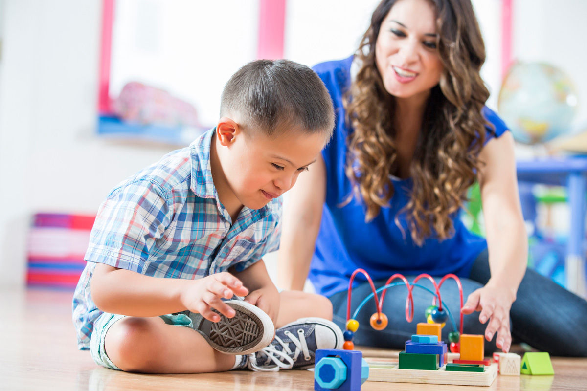Young child sitting on the floor playing with a bead maze toy while a caregiver watches and encourages nearby.